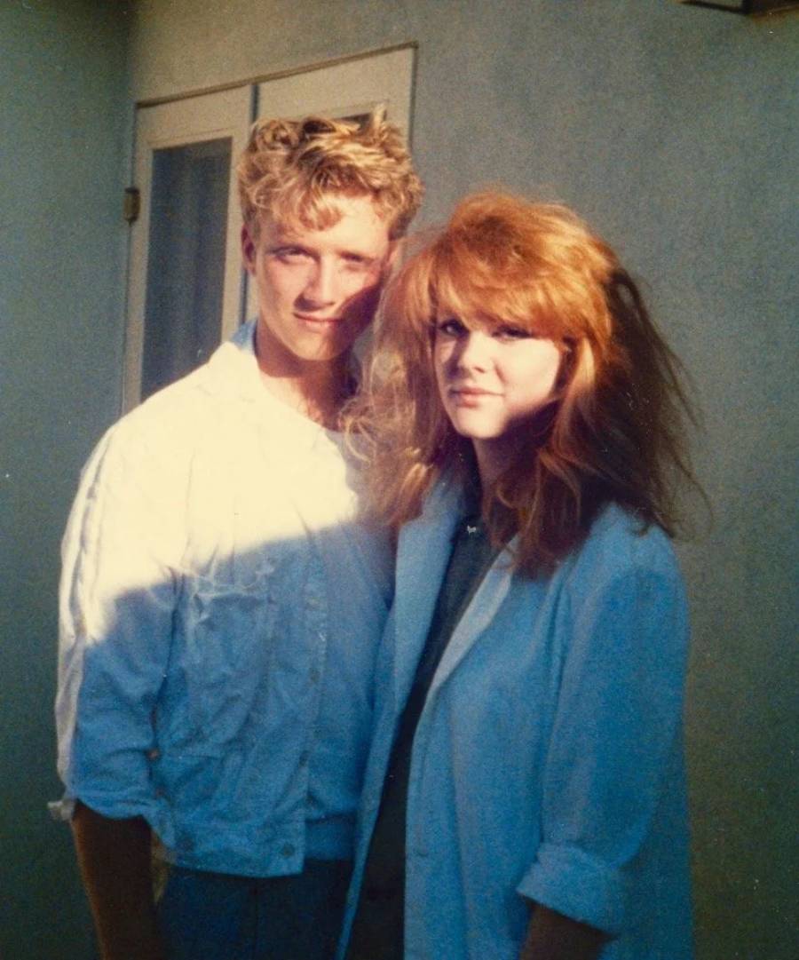 A young man and woman with 1980s-style hair and clothing stand together outside, smiling slightly. Sunlight and shadows fall across them and the light-colored wall behind them.