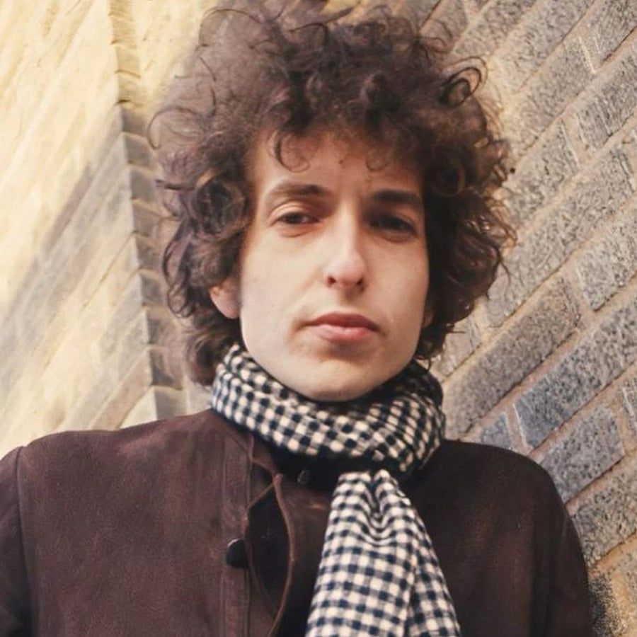 A person with curly hair wearing a checkered scarf and dark jacket stands in front of a brick wall, looking directly at the camera with a neutral expression.