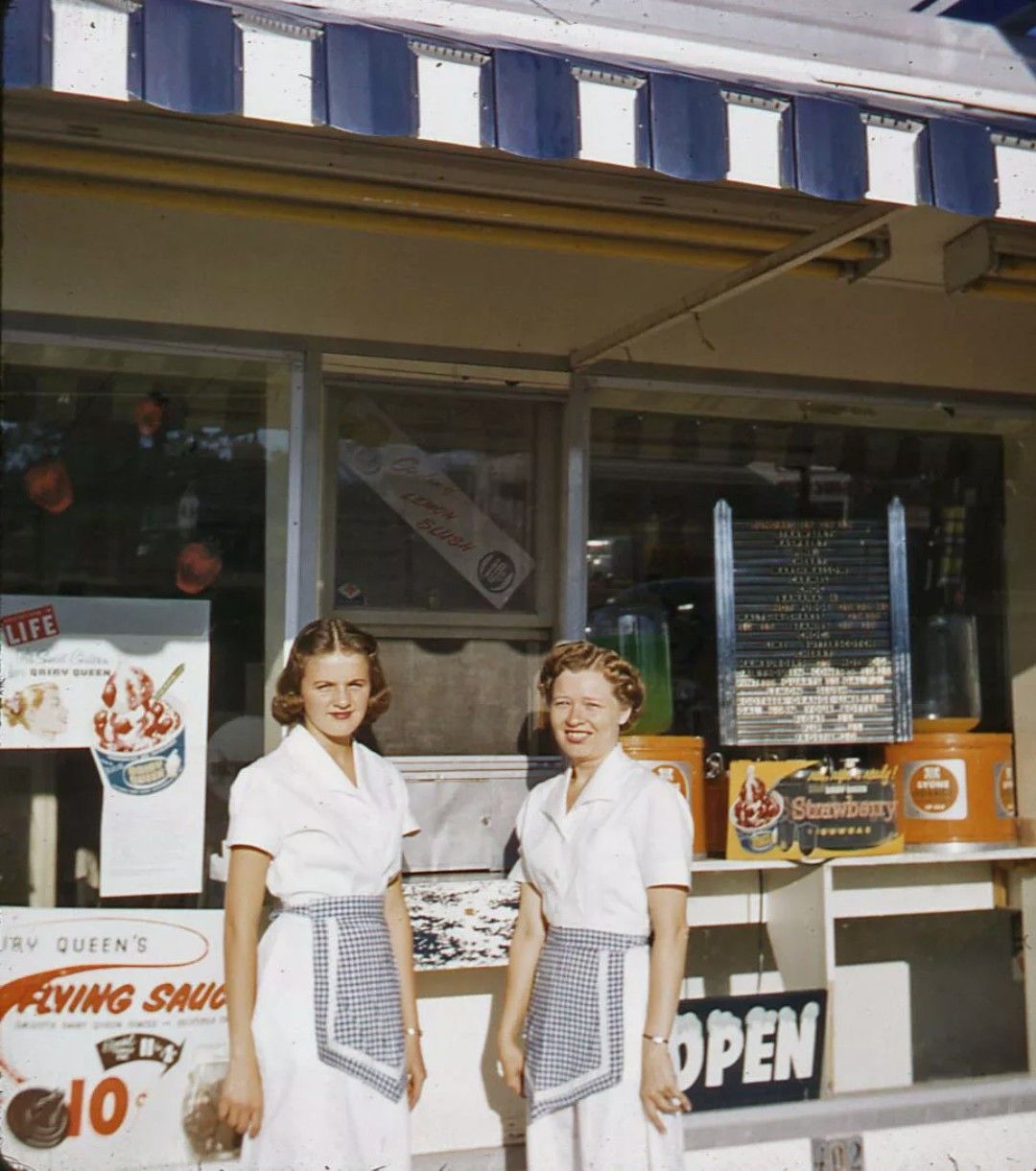 Two women in white uniforms and checkered aprons stand outside an ice cream shop with an "Open" sign, striped awning, and sundae advertisements visible in the background.