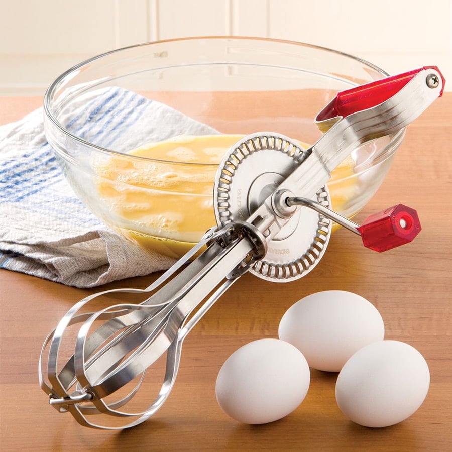 A hand-crank egg beater and three eggs rest on a wooden surface next to a glass bowl of beaten eggs, with a towel in the background.