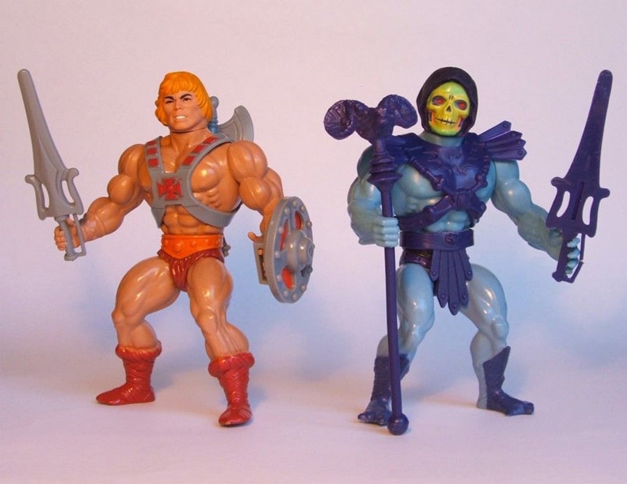 Two action figures stand side by side: one is tan with orange hair, red boots, and a sword; the other is blue with a yellow skull face, dark hood, and armor, holding a staff and sword.
