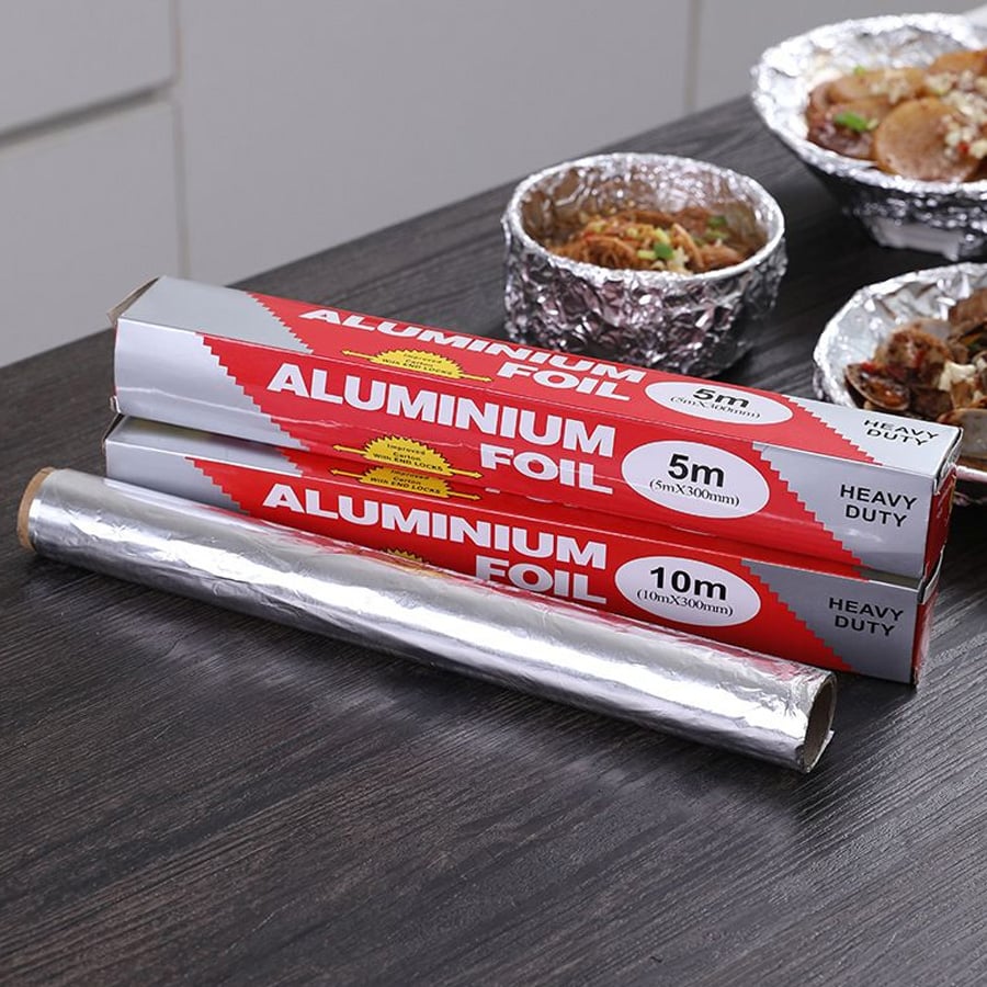 Two boxes and a roll of aluminum foil on a dark counter, with food in foil containers in the background. The boxes are labeled "ALUMINIUM FOIL," "HEAVY DUTY," and show 5m and 10m lengths.