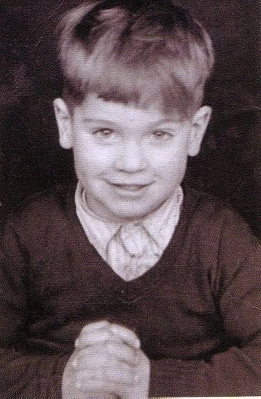 Black and white photo of a young boy with light-colored hair, wearing a collared shirt and dark sweater, smiling slightly while holding his hands together in front of him.