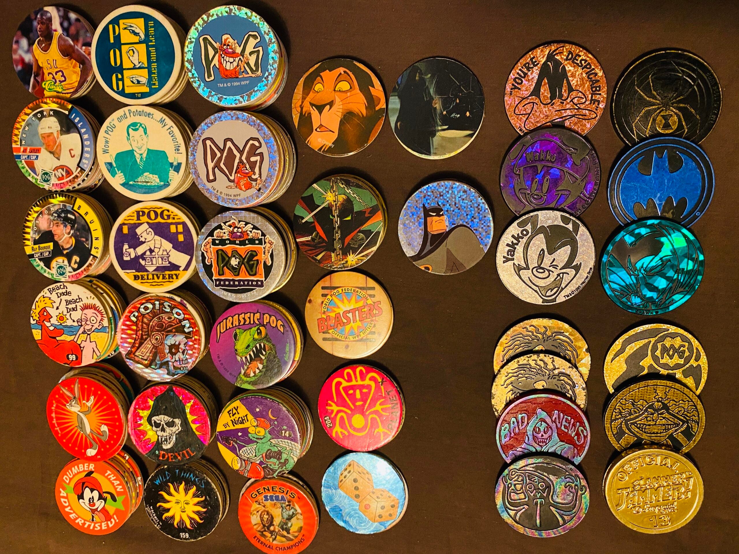 A collection of colorful POG slammers and milk caps with various designs, including cartoon characters, animals, and logos, arranged in rows on a dark surface.