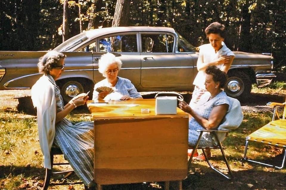 Four women sit outdoors around a table playing cards, with one woman standing nearby. A vintage car is parked in the background among trees on a sunny day.