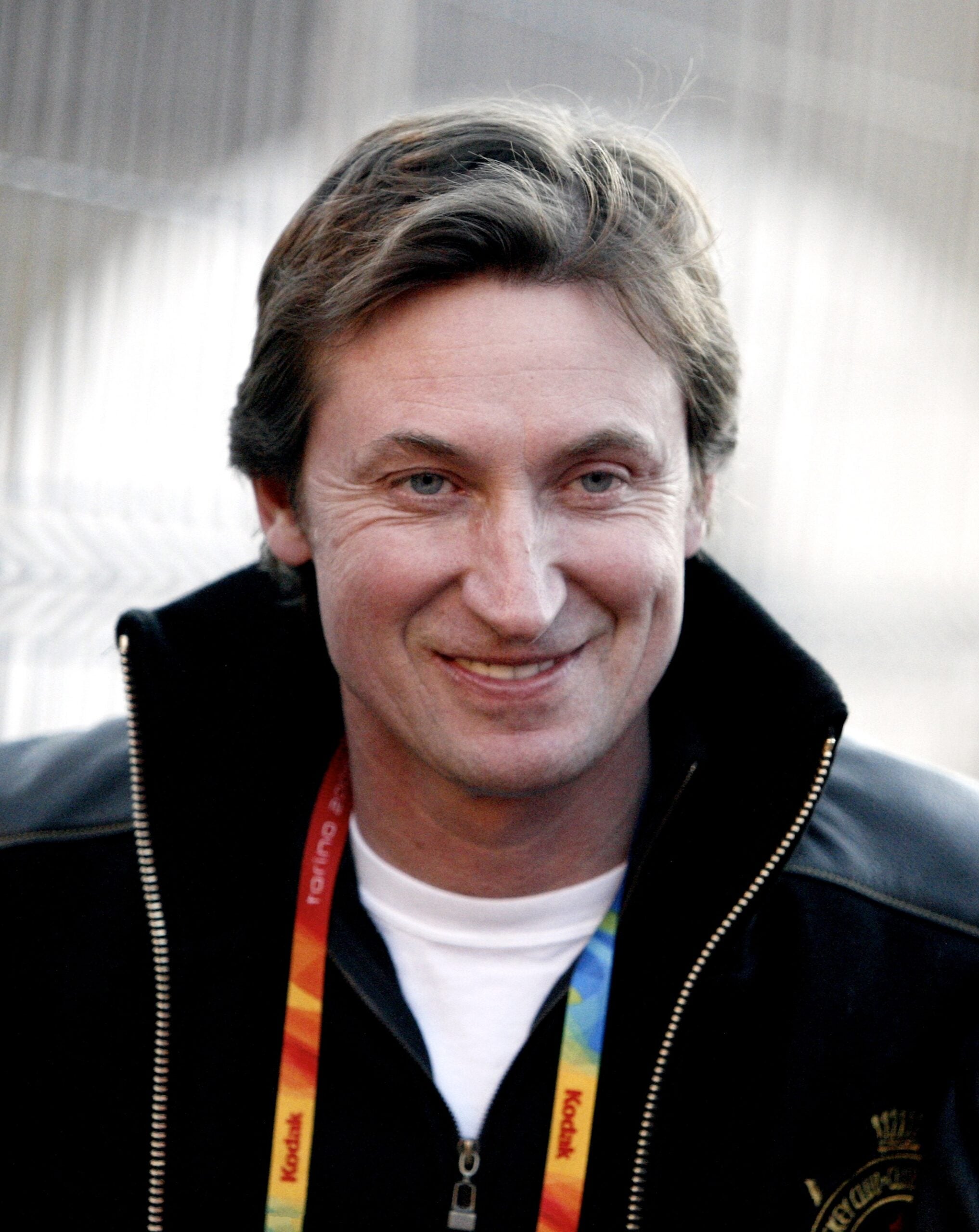 A man with light brown hair wearing a black jacket and a colorful lanyard around his neck, smiling in an outdoor setting.