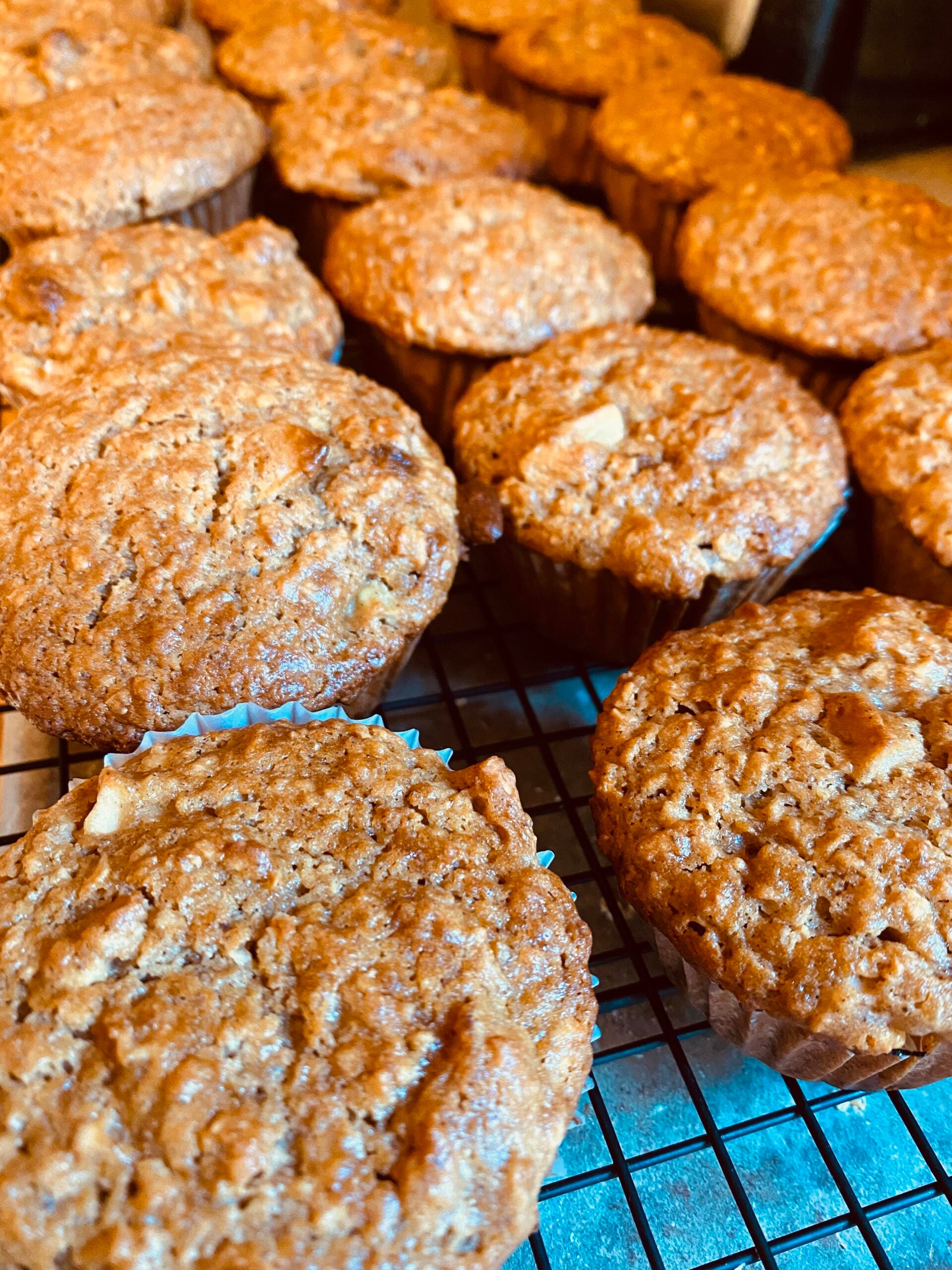 A close-up of freshly baked muffins cooling on a black wire rack. The muffins are golden brown with a slightly cracked surface, showing a homemade texture.