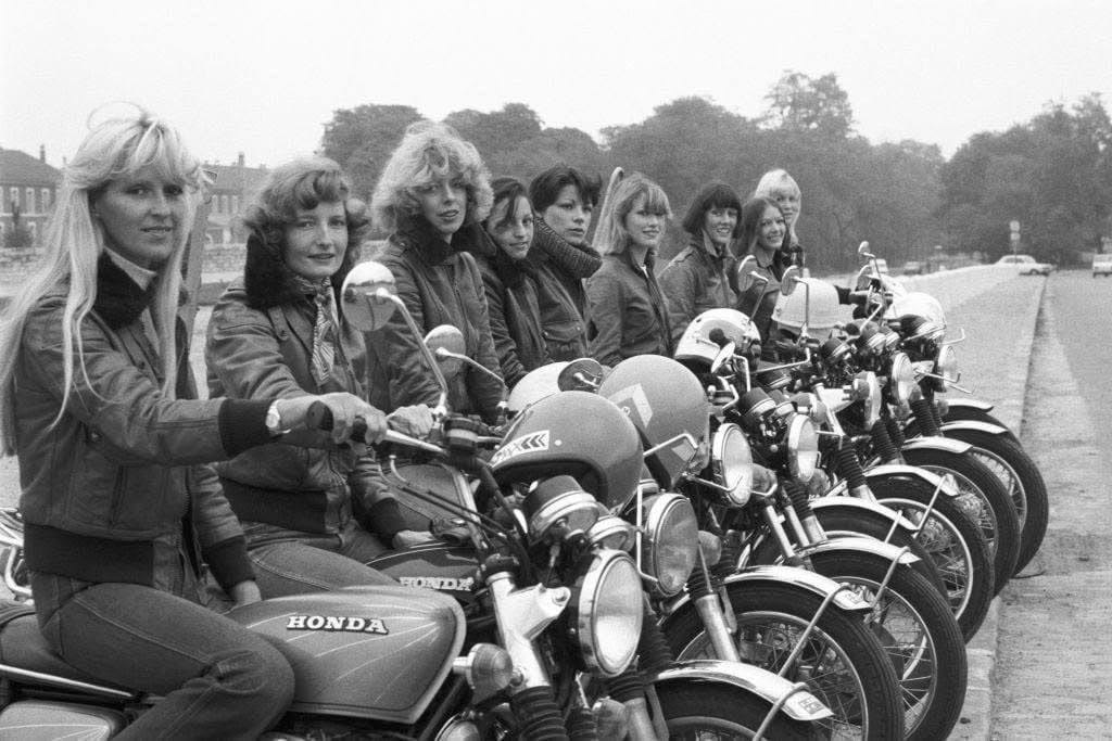 A row of women sit on motorcycles parked along a curb, wearing jackets and looking toward the camera. The bikes are mostly Honda models. Trees and buildings are visible in the background.