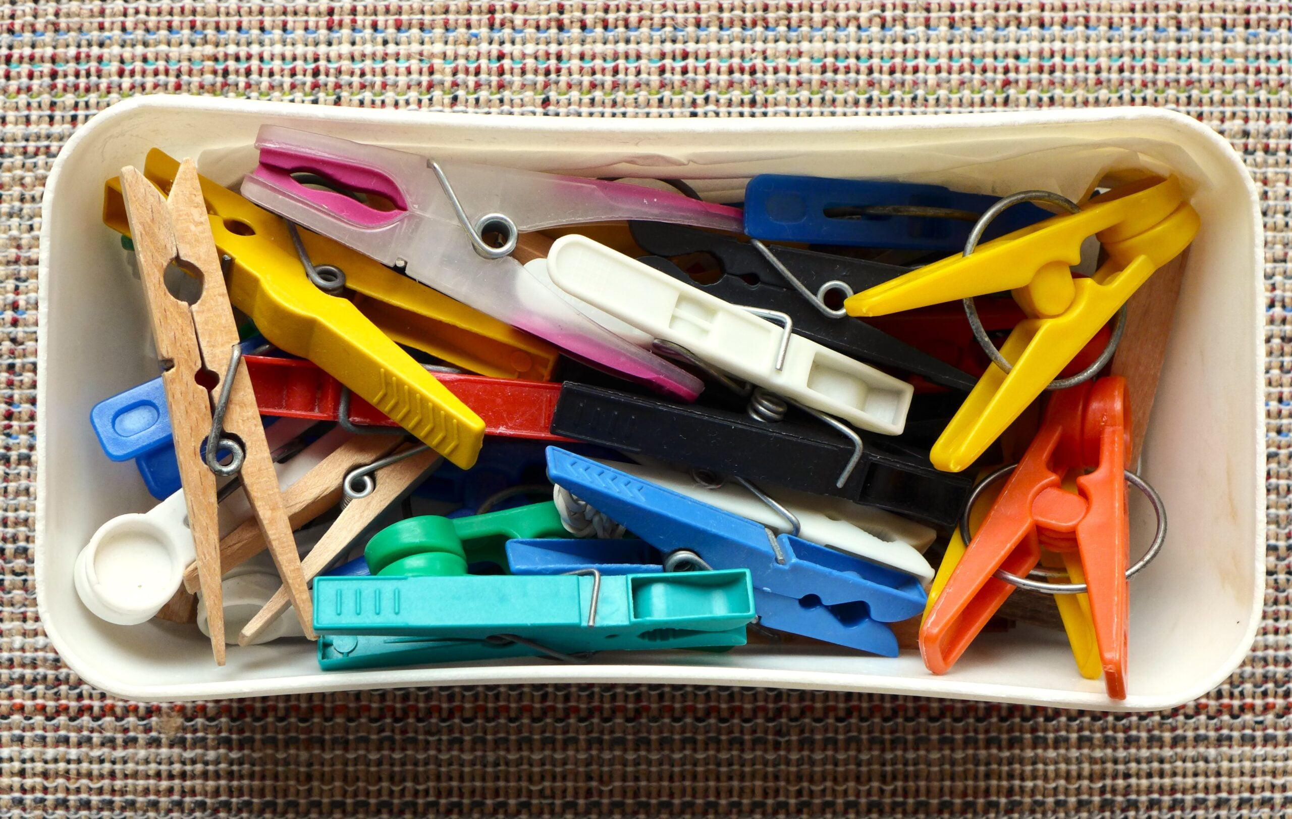 A white container filled with assorted colorful clothespins, including wooden and plastic types, placed on a textured fabric surface.