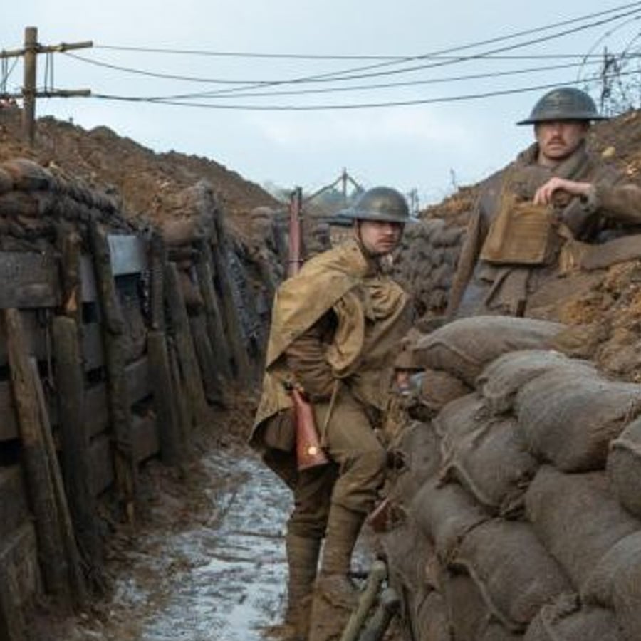 Two soldiers in World War I uniforms stand in a muddy trench lined with sandbags and wooden planks, with barbed wire and utility lines visible above them under a cloudy sky.