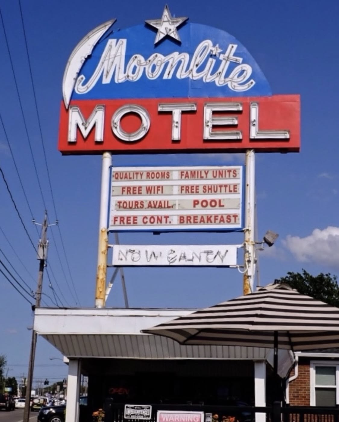 Retro "Moonlite Motel" sign with neon lights above a small motel. The sign advertises free WiFi, shuttle, tours, pool, and breakfast. Blue sky, power lines, and patio umbrella are visible.