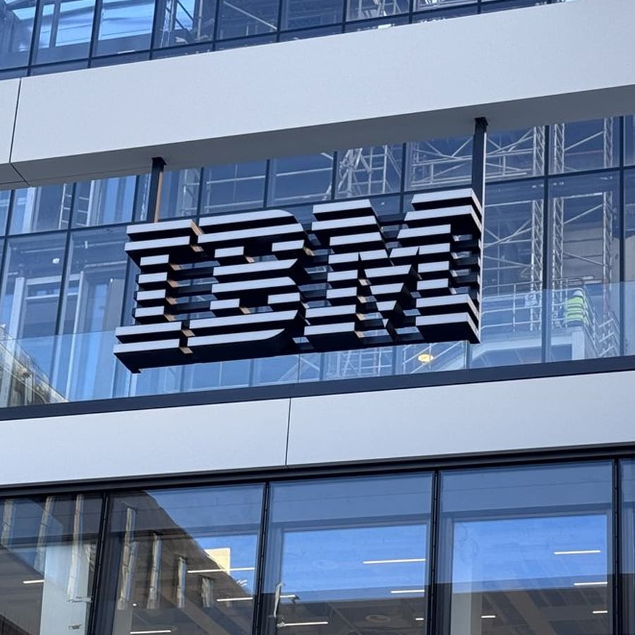 A large, striped IBM logo is mounted on the glass exterior of a modern office building with reflective windows.