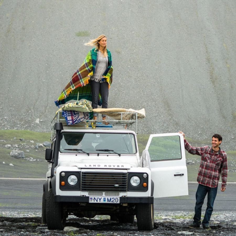 A woman stands on top of a white Land Rover covered in blankets and bags, while a man in a plaid shirt stands by the open driver's door, outdoors with a grassy, rocky background.