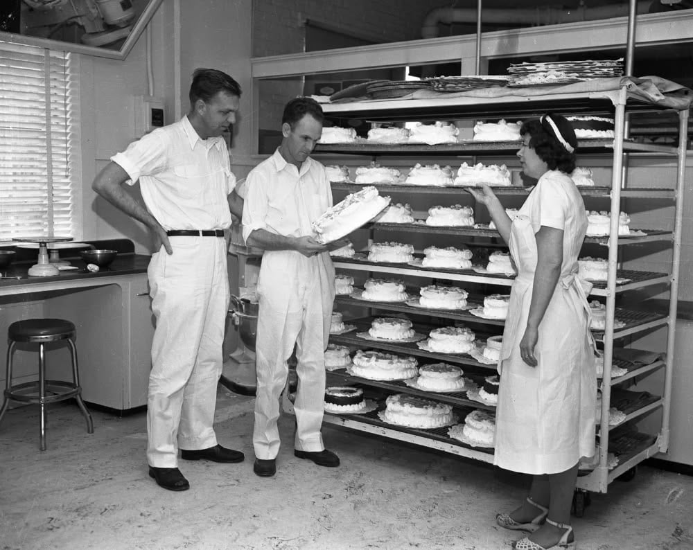 Three people in white uniforms stand by large racks filled with pies in a bakery kitchen. One man examines a pie while the others look on.