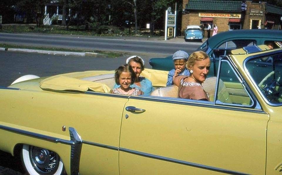 A woman and three children sit in a yellow convertible car parked outside, all looking toward the camera. A store and other cars are visible in the background.
