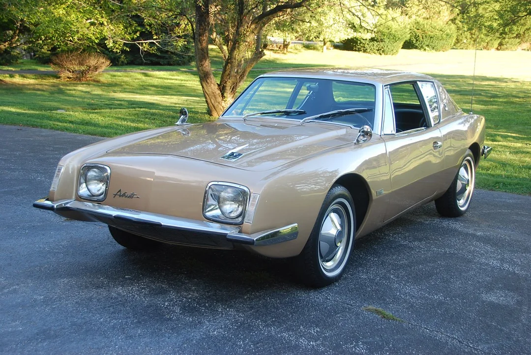 A beige vintage Studebaker Avanti car is parked on a driveway surrounded by grass and trees on a sunny day.