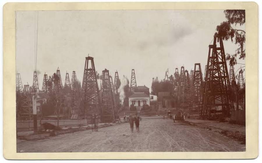 Sepia-toned photo of an early oil field with numerous wooden derricks, a few buildings, and two people walking on a dirt road in the foreground. Trees are visible around the oil field.