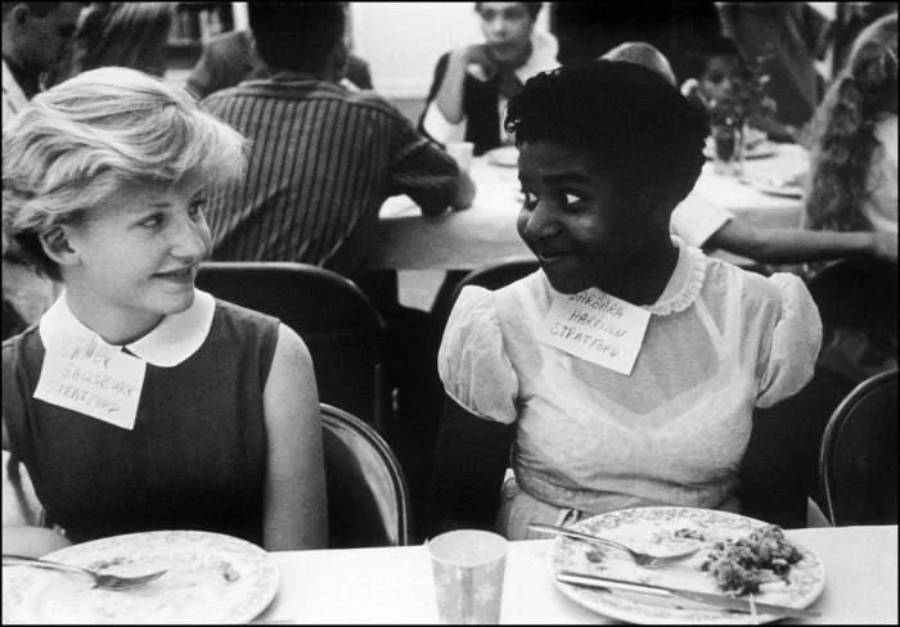 Two young girls, one white and one Black, sit side by side at a lunch table, smiling at each other. Both wear name tags, and plates of food are in front of them. Other children are visible in the background.