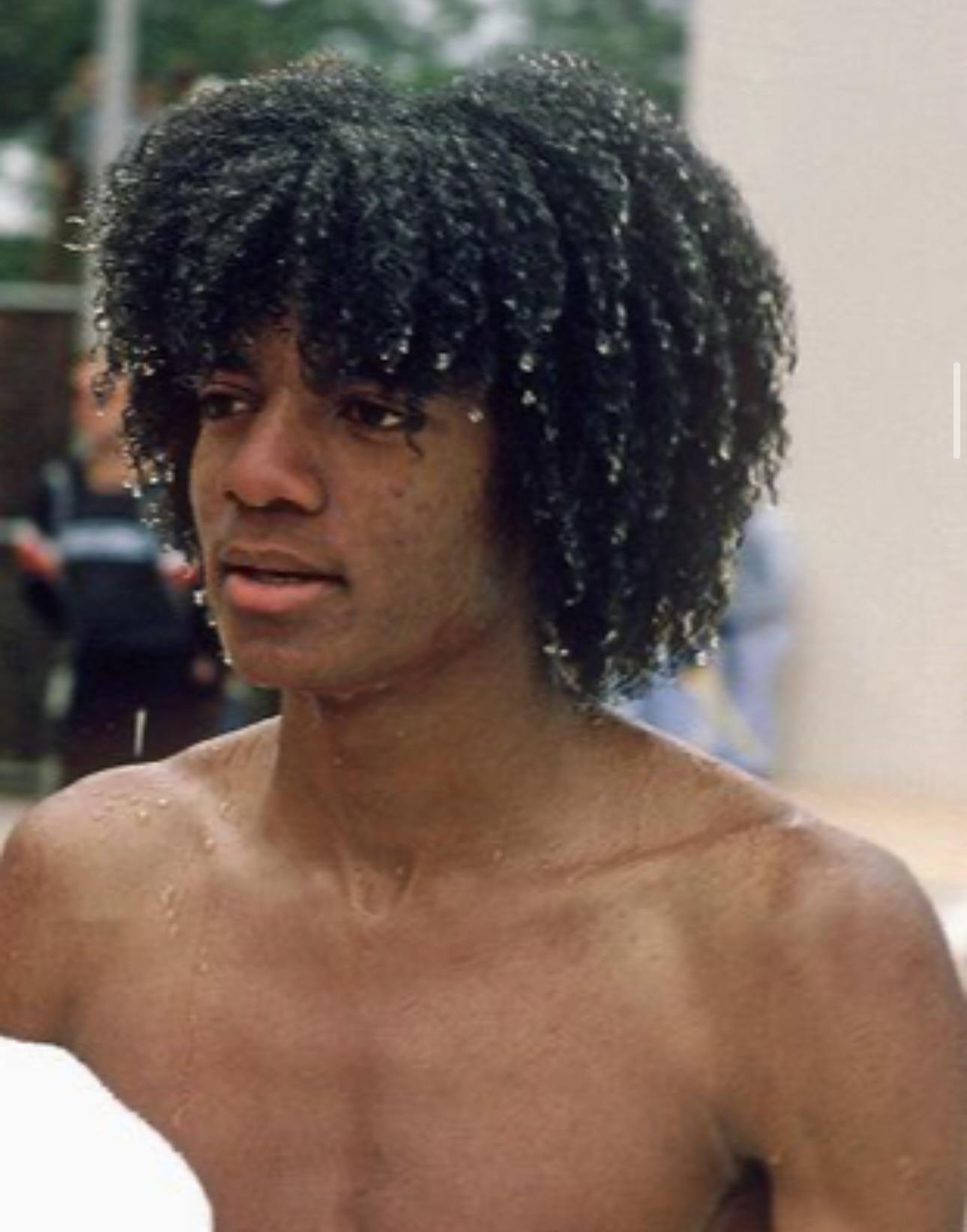 A young person with curly wet hair stands shirtless outdoors, water droplets on their skin and hair. The background is blurred, suggesting a pool or outdoor setting.