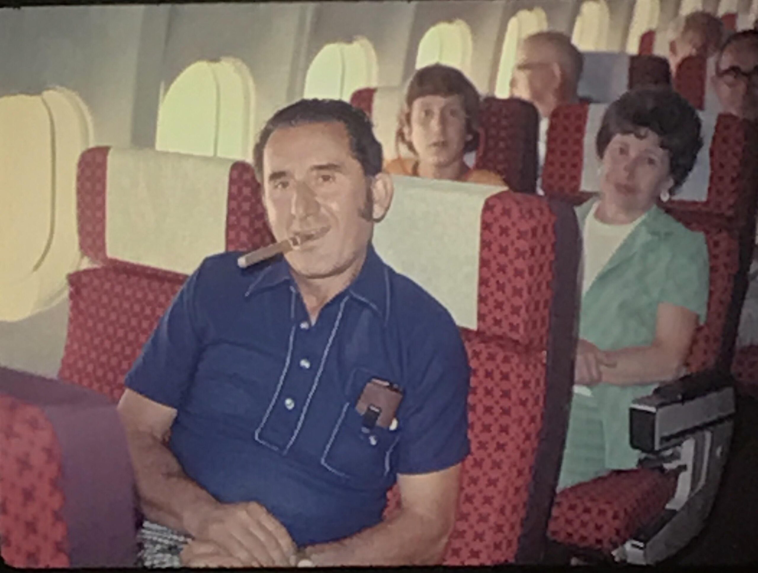 A man sits on an airplane with a cigar in his mouth, wearing a blue shirt. Passengers are seated behind him, and sunlight streams through the windows.