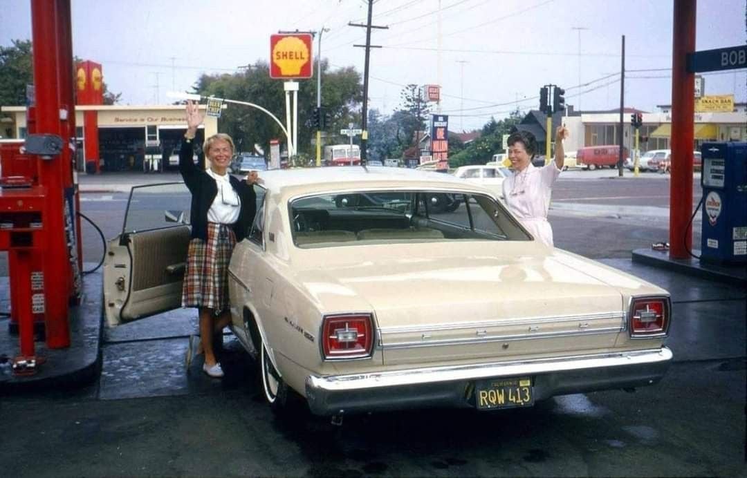 Two women stand by a cream-colored vintage car at a gas station. One woman waves while standing by the open driver's door. The other stands near a gas pump. A Shell station and street are visible in the background.