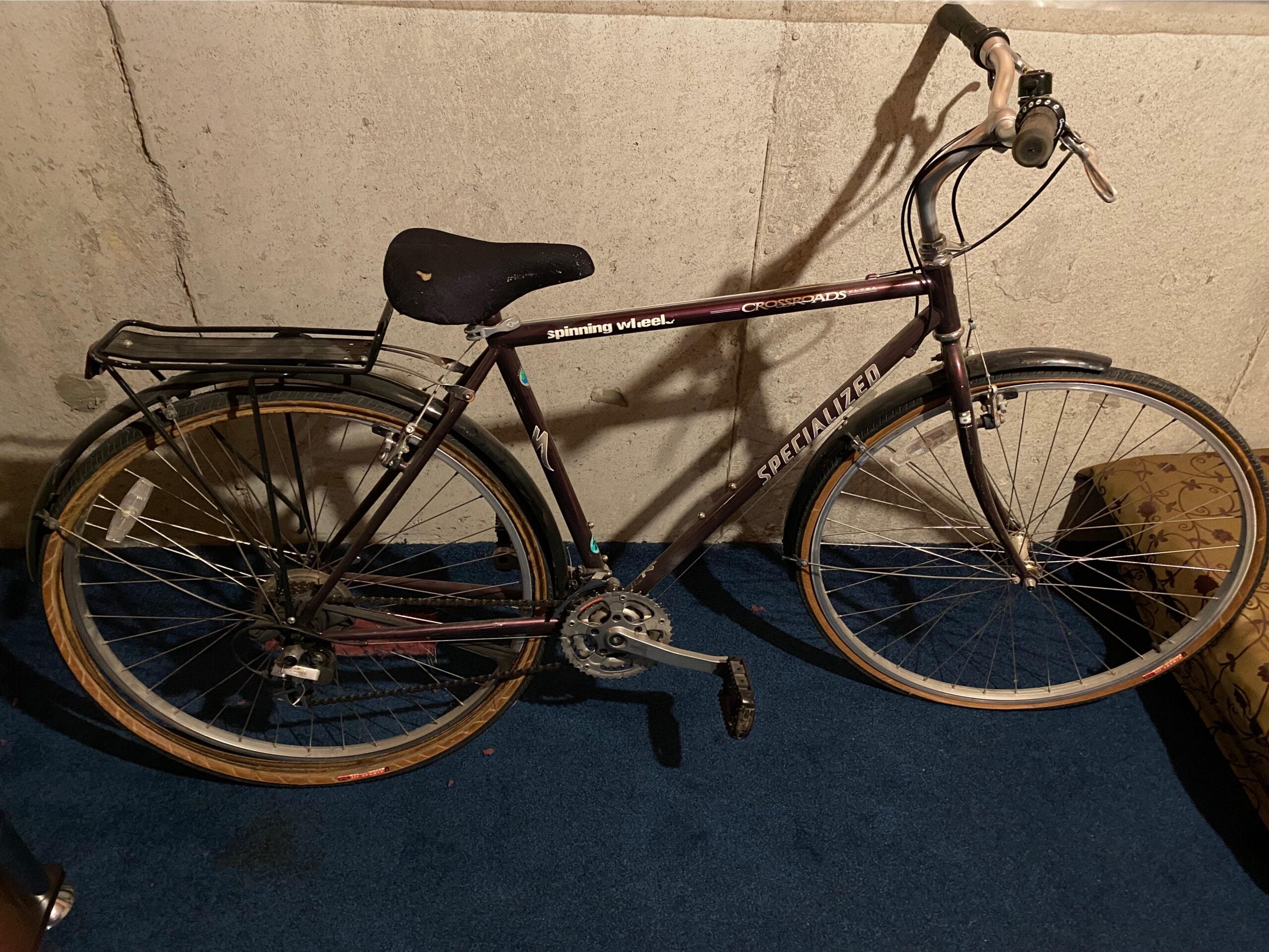 A vintage Specialized Crossroads bicycle with a black seat and rear rack, resting on a blue carpet against a concrete wall in a dimly lit room.