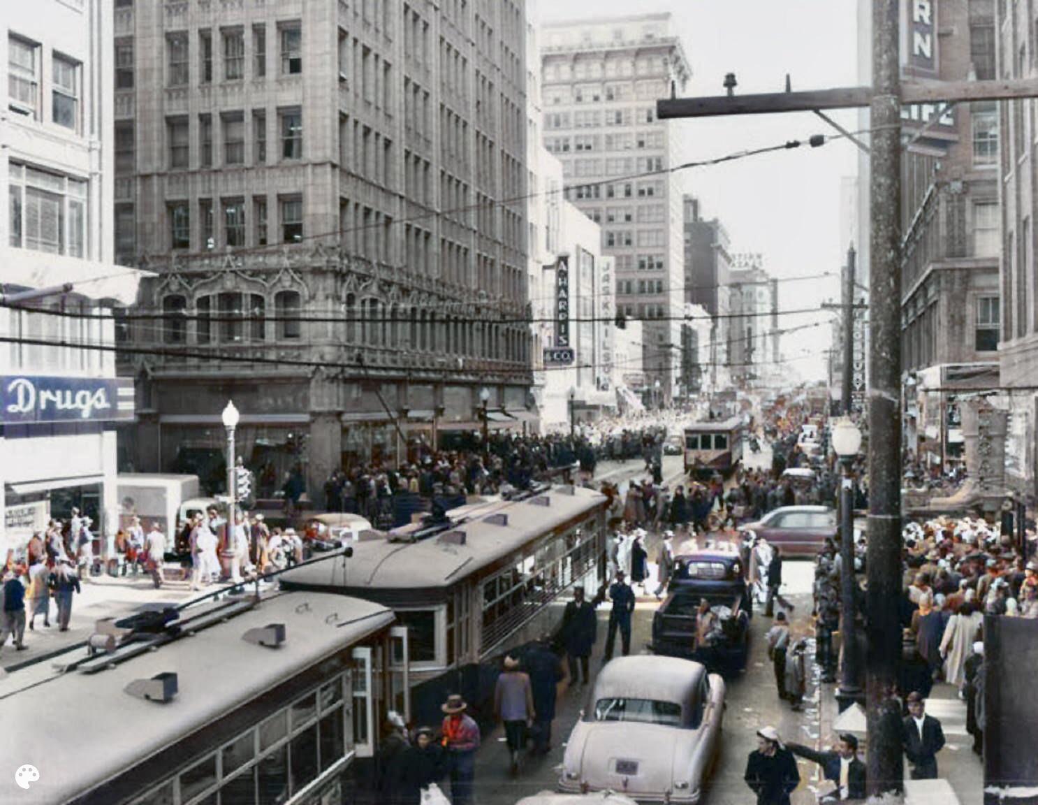 A busy city street scene from the mid-20th century, with crowded sidewalks, vintage cars, streetcars, and tall buildings lining both sides of the street. People are gathered and moving throughout the area.