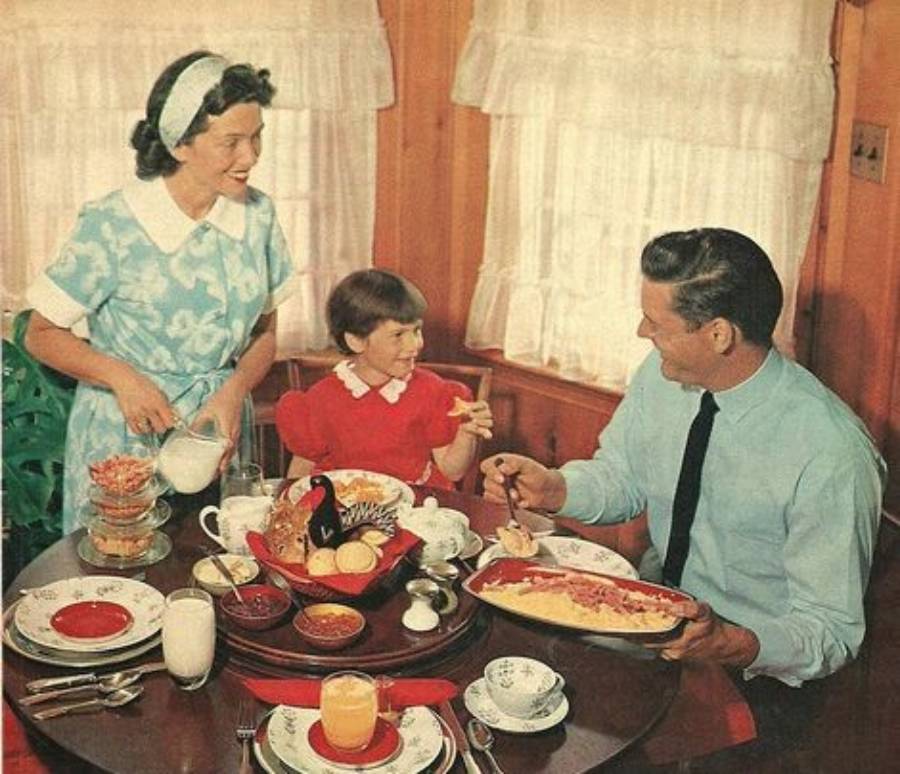A woman in a blue dress pours milk as a man in a tie and a smiling young girl in red sit at a table set with breakfast dishes in a cozy, wood-paneled room with lace curtains.