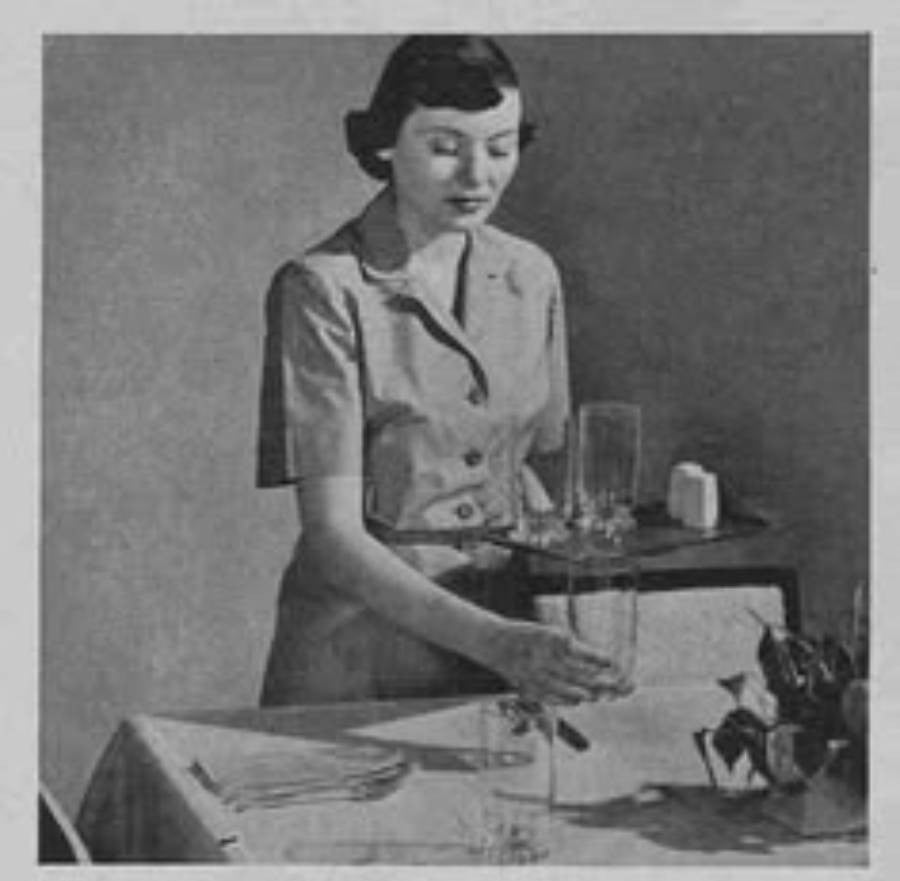 A woman in a vintage dress clears empty glasses from a table, holding a tray. The table has a napkin and a small plant. The image is in black and white.