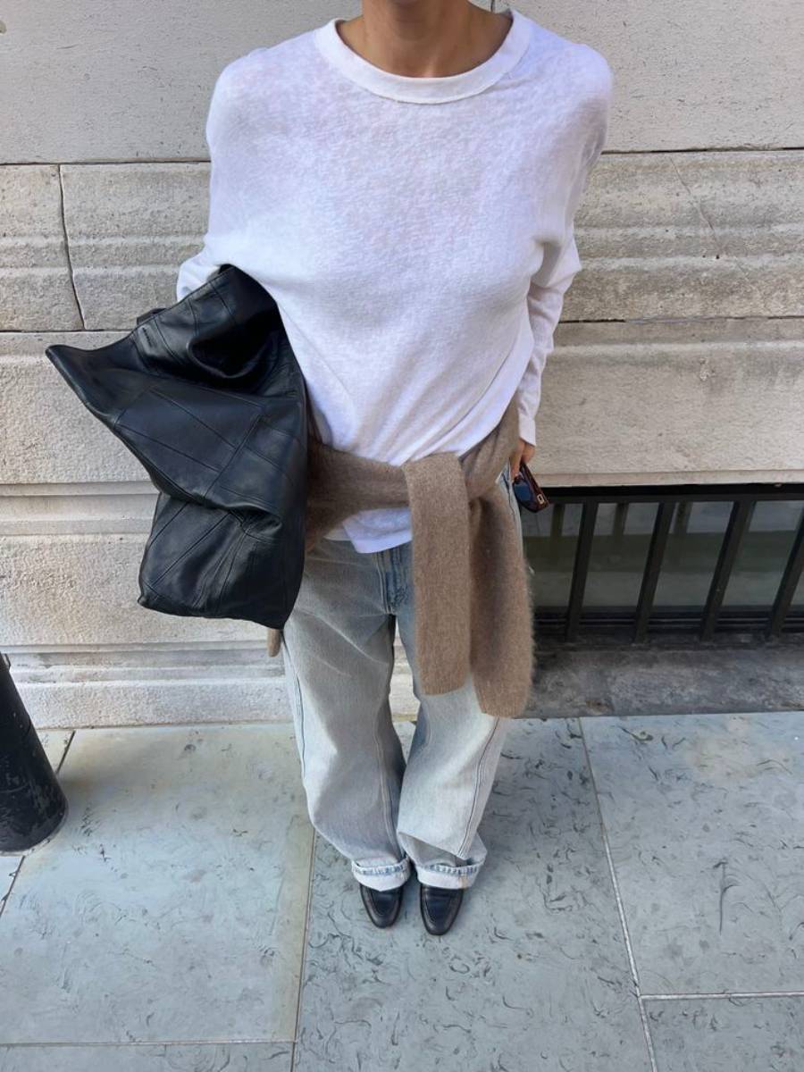 Person wearing a sheer white long-sleeve shirt, light blue jeans, black shoes, and a brown sweater tied at the waist. They are holding a black jacket, standing on a stone pavement in front of a beige wall.