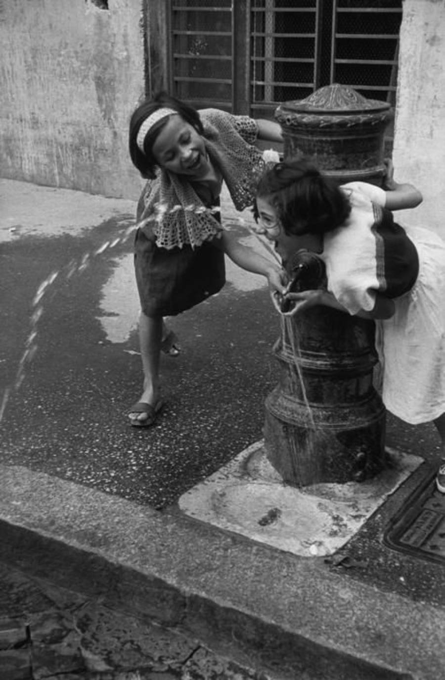 Two young girls share a laugh as they drink water from a public fountain on a city street, with water splashing around them.