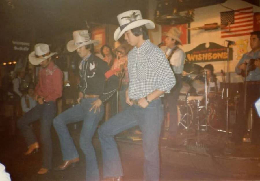 Four people in cowboy hats and boots line dancing on stage while a band plays in the background. The scene is indoors with American and Mexican flags on the wall. The lighting is warm and the atmosphere appears lively.