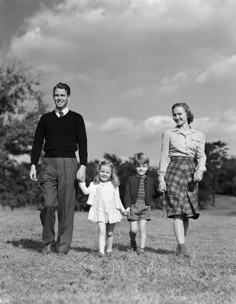 A man, woman, and two young children are walking hand in hand in a grassy field under a partly cloudy sky, all smiling and dressed in 1940s-style clothing.