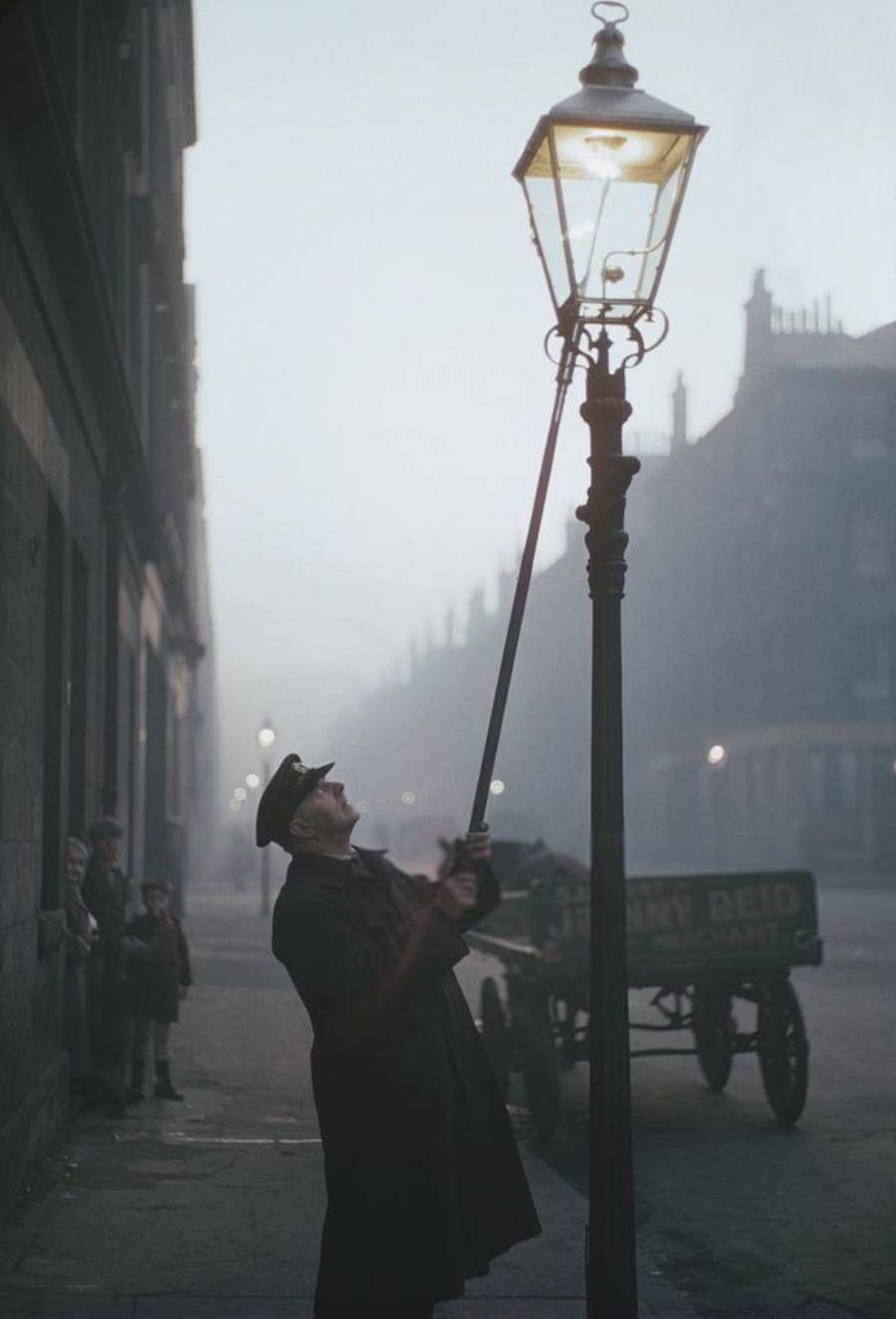 An older man in a long coat and cap uses a pole to light a streetlamp on a foggy city street, with a horse-drawn cart and several children watching in the background.