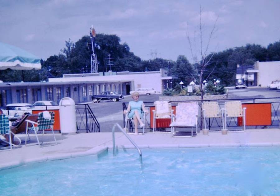 A woman sits on a chair by a motel pool with retro lounge chairs around her. Classic cars and a motel building are visible in the background under a partly cloudy sky.