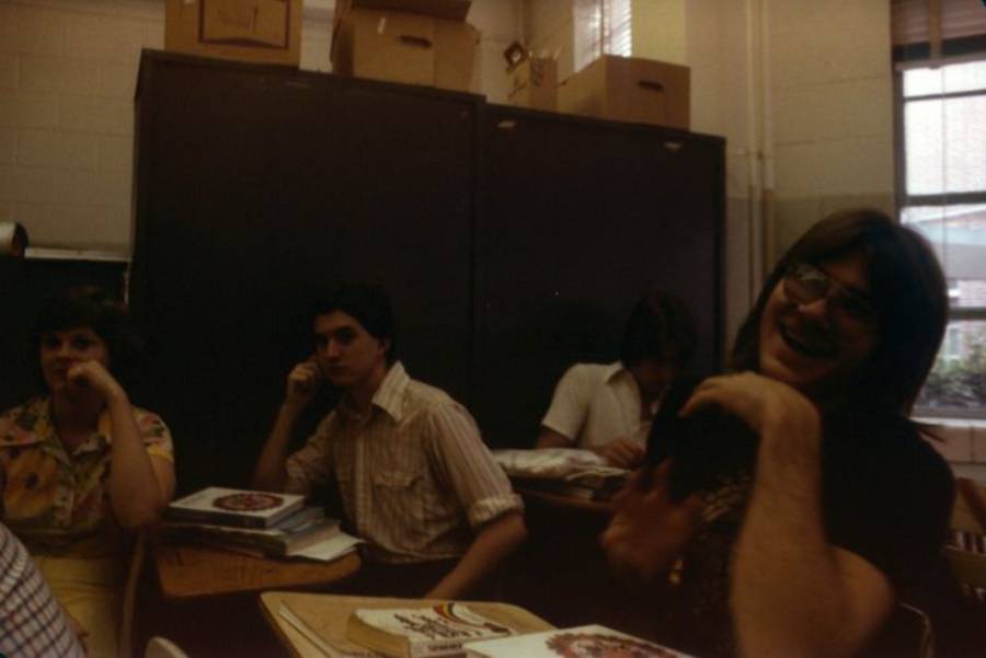 Four students sit at desks in a dimly lit classroom with books and papers. One student in the front right is laughing, while others appear to be listening or engaged with something off-camera. Boxes are stacked on top of cabinets in the back.