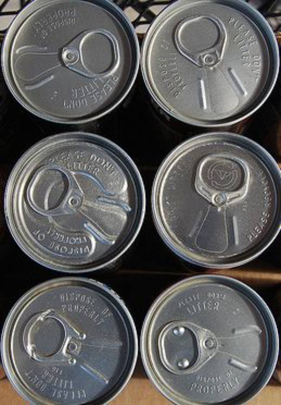 Six unopened aluminum cans are arranged in two rows of three, viewed from above. The can tops have pull tabs, and some text is visible on the lids. Lighting creates slight shadows and highlights on the metal surfaces.