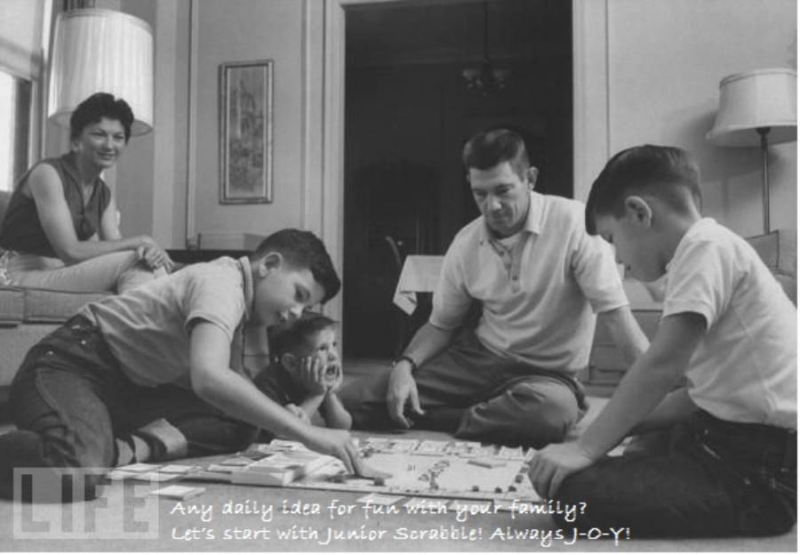 A family of five sits on the floor in their living room, smiling and playing Junior Scrabble together. The room looks cozy and inviting, with a couch, lamp, and coffee table in the background.