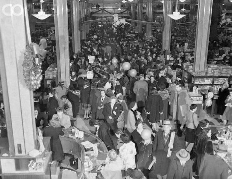 Black and white photo of a crowded department store during a holiday sale, with shoppers wearing coats and hats, standing in long lines and browsing counters filled with goods. Holiday decorations are visible above the crowd.