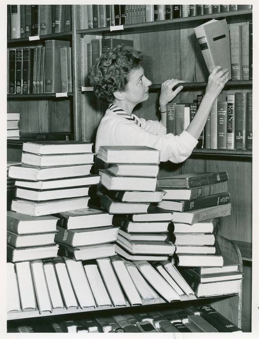 A woman takes a book from a high shelf in a library. She stands behind a table stacked with many books, reaching up with one hand while holding another book. Shelves filled with books cover the background.