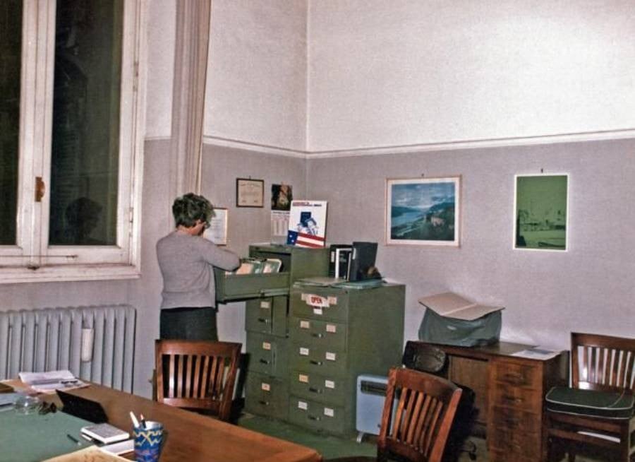 A person stands by a green filing cabinet in an office with several chairs, a table, and papers. The walls have a few framed pictures, and there is a window with white trim on the left.