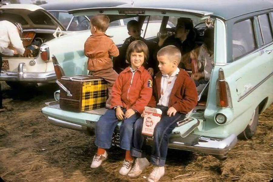 Two children sit on the open tailgate of a vintage pale blue station wagon, with a picnic basket beside them. Other kids and adults are in or near the car. The scene appears to be at an outdoor gathering or picnic.