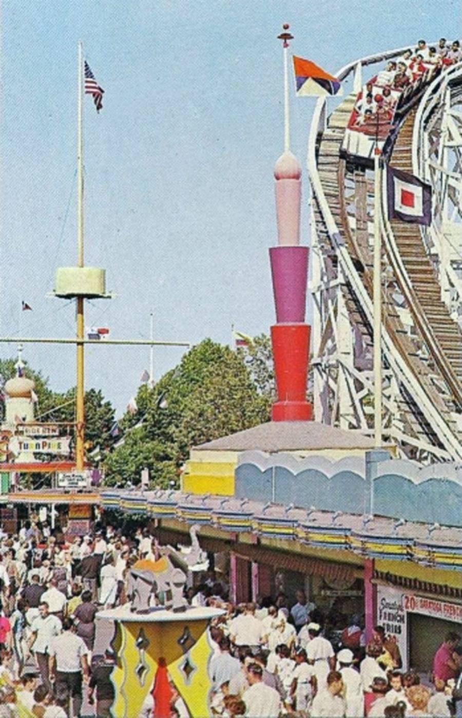 A crowded amusement park scene with people walking below a colorful building, while a roller coaster with riders in white cars ascends a wooden track in the background under a clear sky.