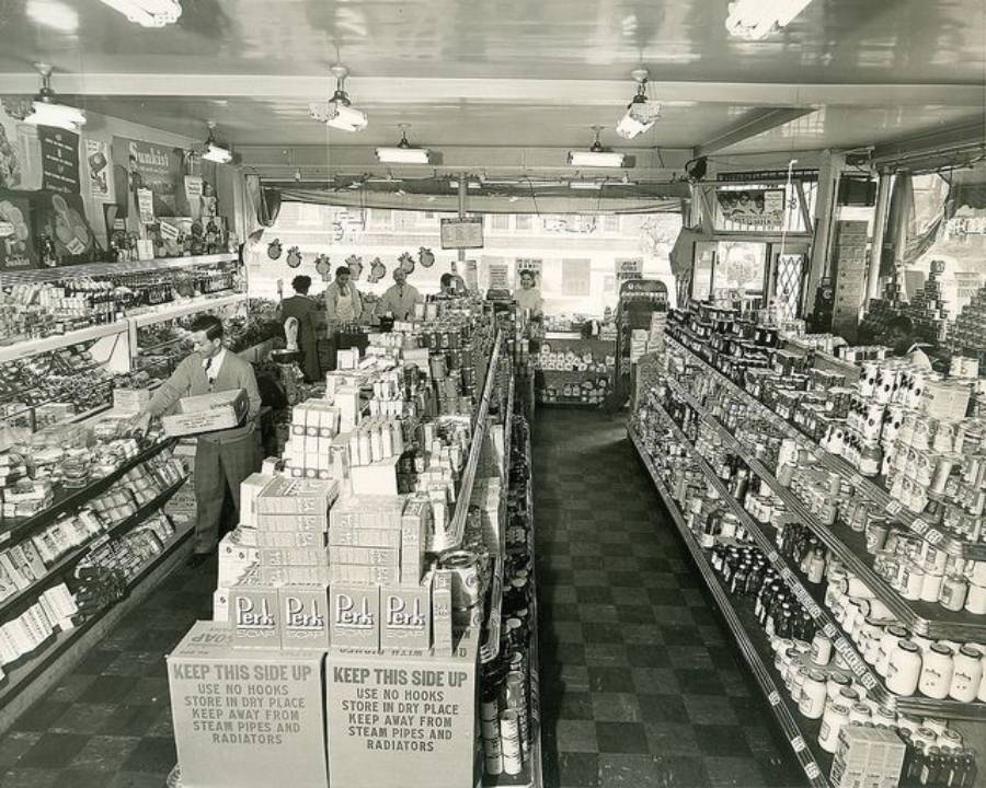 Black and white photo of a vintage grocery store interior with shelves stocked with canned goods and packaged products. Several people, including staff, are shopping or working near the checkout counter.