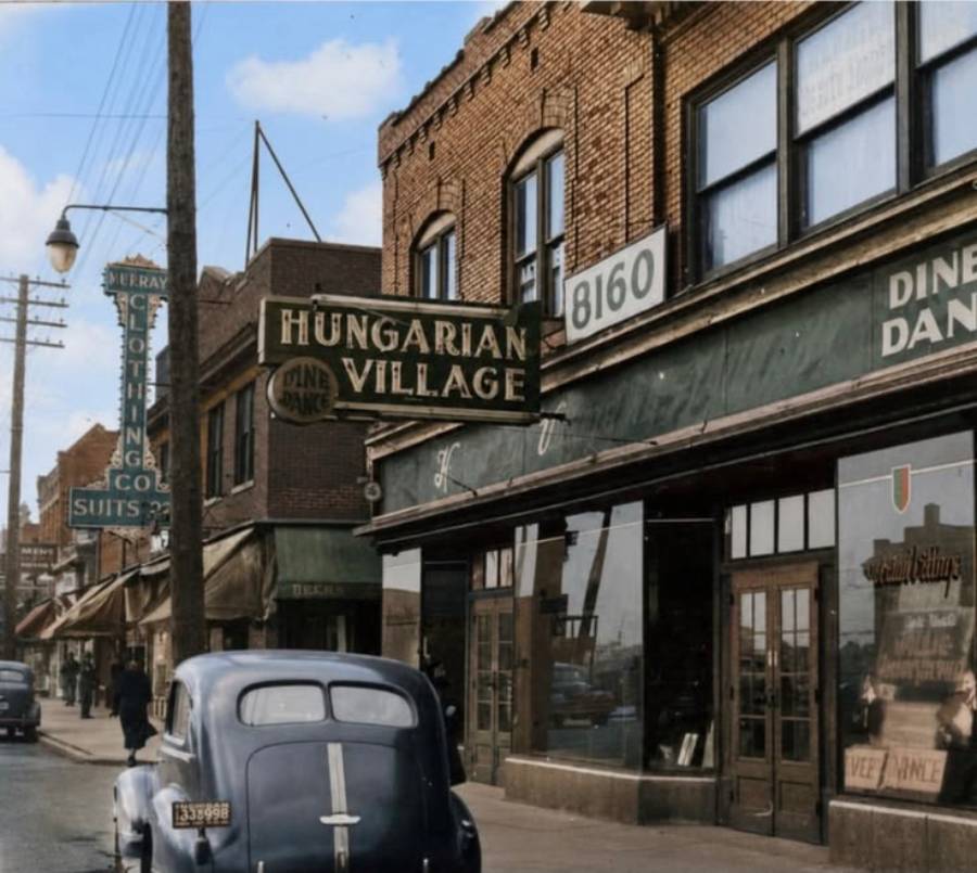A vintage street scene features brick buildings, a "Hungarian Village" sign, storefronts, and a parked 1940s car. Businesses include a diner and a clothing store. The image has a nostalgic, early 20th-century feel.