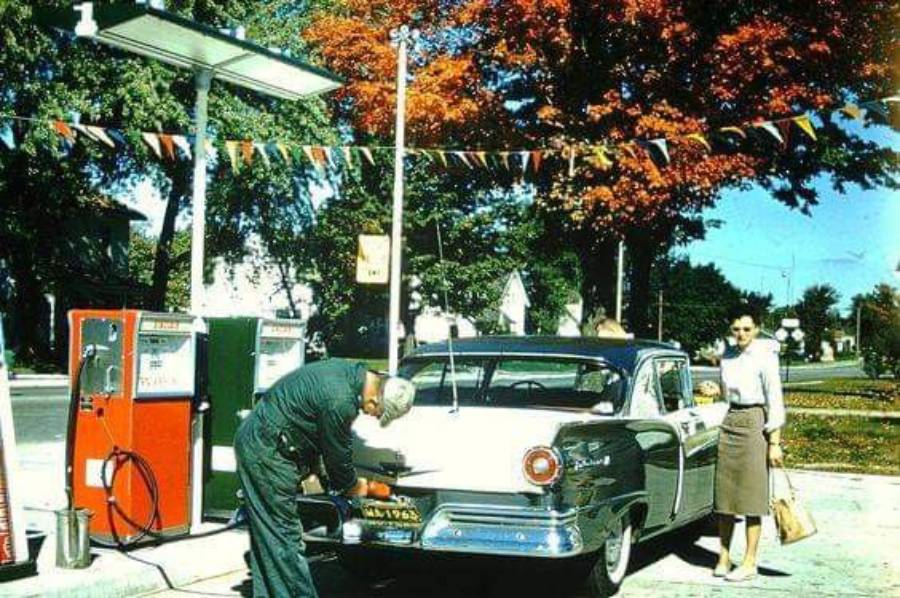 A gas station attendant fills up a classic car at a vintage gas station while a woman stands nearby. The scene is bright and colorful with autumn trees and festive pennant flags overhead.