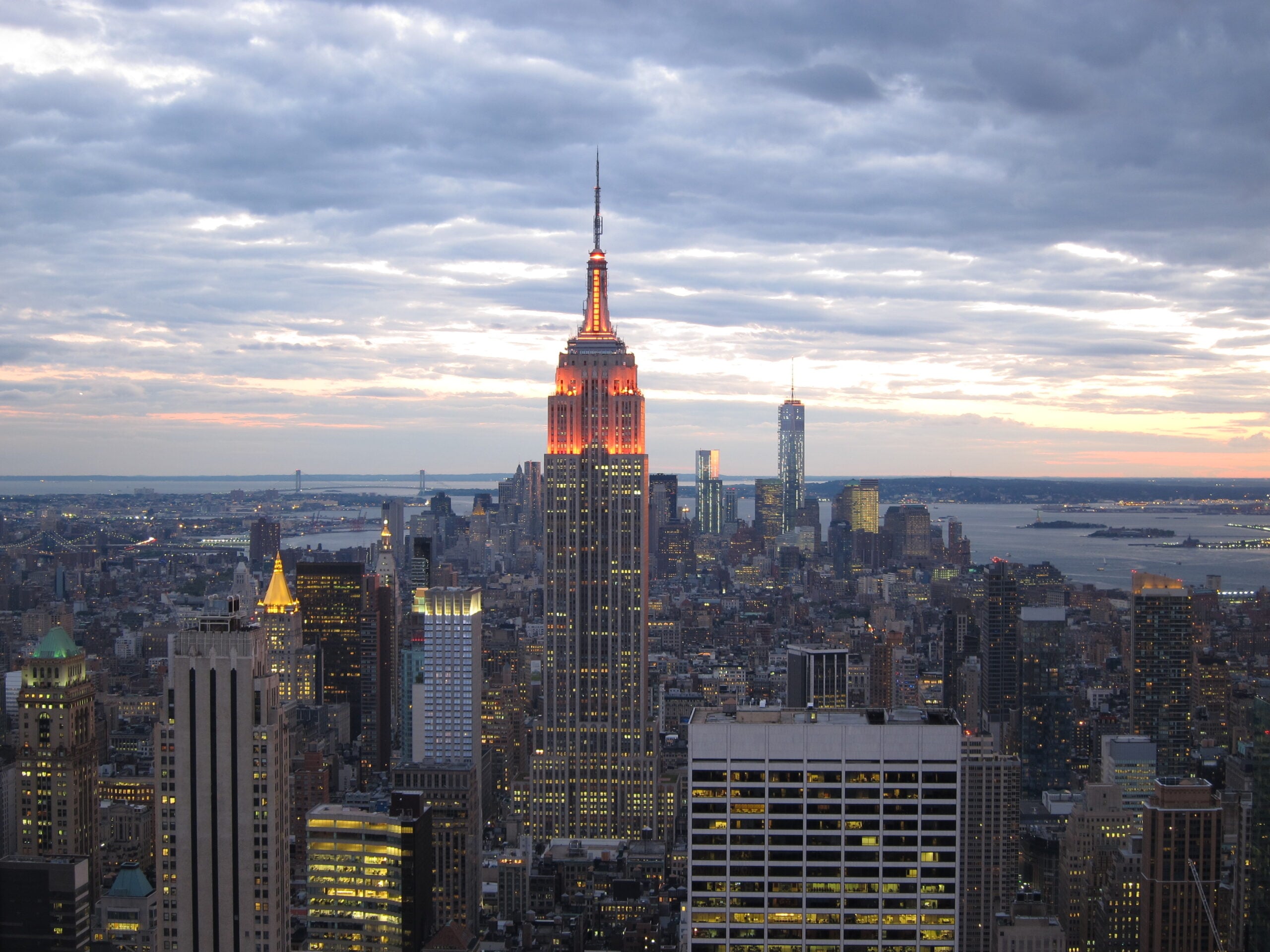 The Empire State Building lit up at dusk, surrounded by other skyscrapers in New York City, with a cloudy sky and the Hudson River in the background.