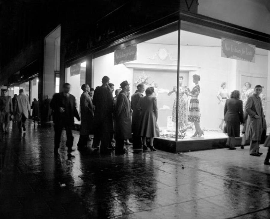 A group of people stands outside a brightly lit shop window at night, looking at mannequins dressed in elegant clothing. The wet pavement reflects lights, and pedestrians walk along the sidewalk in coats and hats.