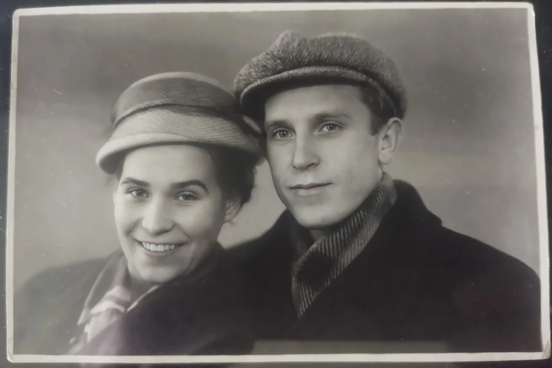 A vintage black-and-white portrait of a smiling woman and a man in hats and coats, posing closely together and looking at the camera against a plain background.