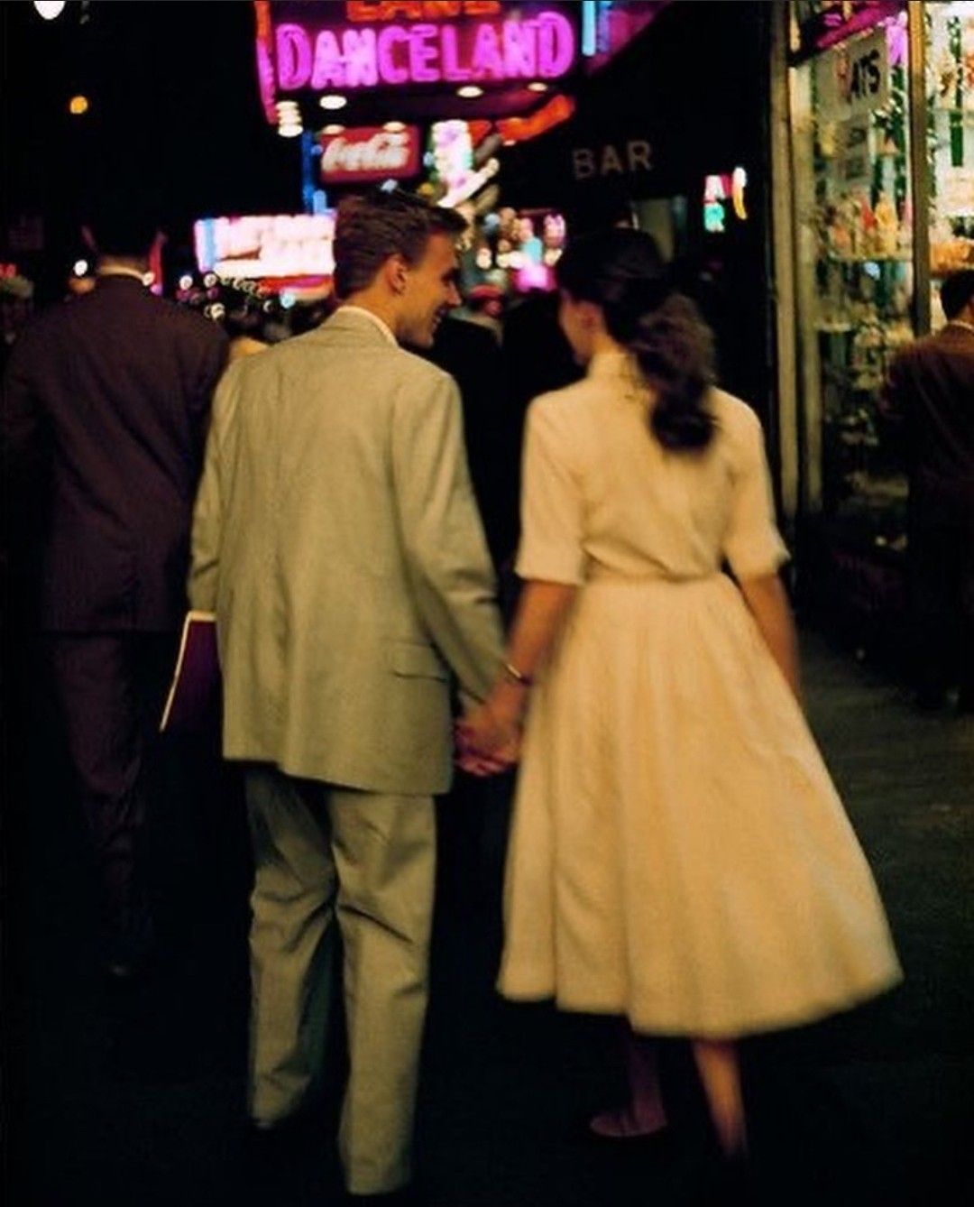 A couple dressed in vintage clothing holds hands while walking down a neon-lit street at night. Bright signs, including one reading “DANCELAND,” illuminate the lively urban scene.